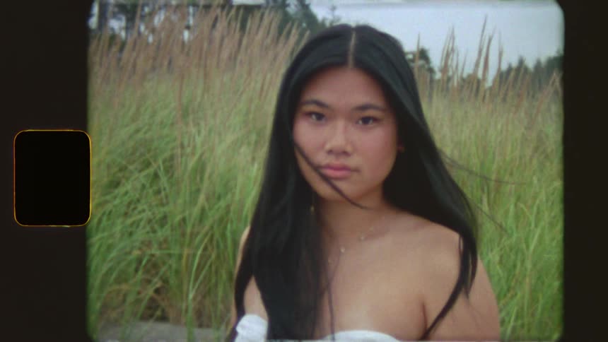 Vintage film portrait of a serious young woman with long black hair standing in a field of tall grass