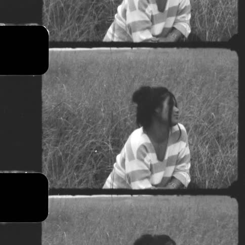 A young woman in a striped shirt sits peacefully in a field of tall grass in this vintage, black and white film-style video