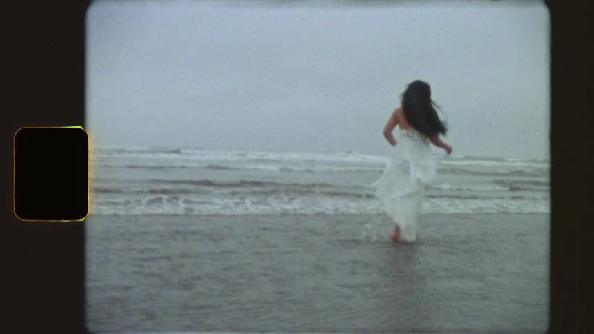 A carefree woman in a flowing white dress runs and plays in the shallow ocean waves on an overcast day in this vintage-style footage