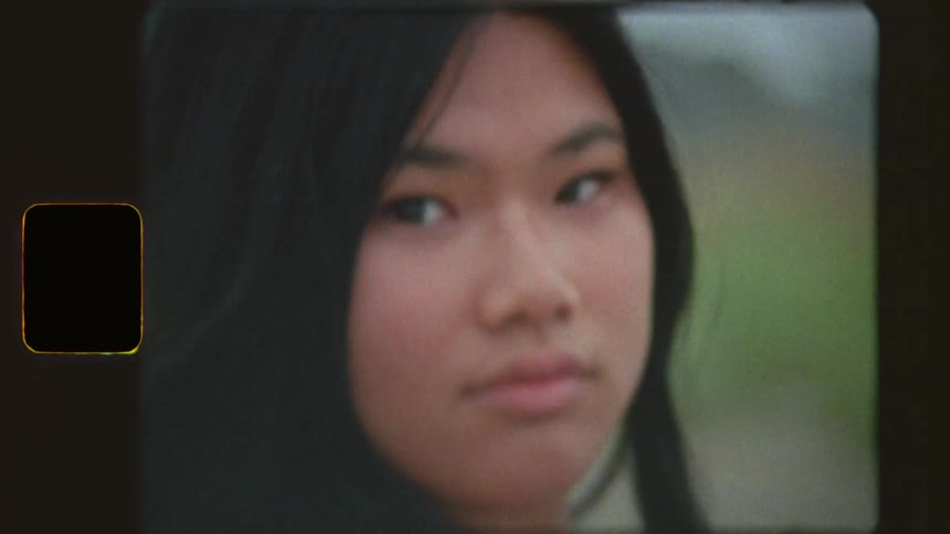 Vintage film footage captures a pensive young woman with long hair in a white dress standing on a grassy beach, ending with a close-up of her face