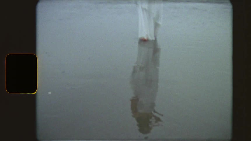 Vintage 8mm film captures a solitary woman in a long white dress walking along the shoreline of a moody, overcast beach