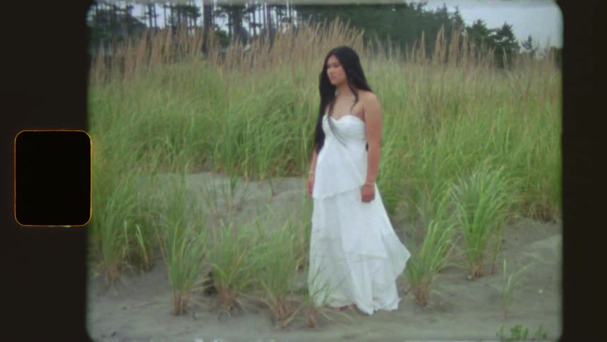 A woman in a flowing white dress stands serenely among tall grass on a sandy beach in this vintage-style footage