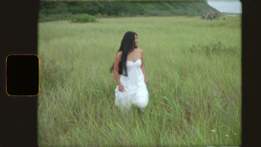Dreamy, vintage-style footage captures a young woman in a white dress walking slowly through a field of tall green grass