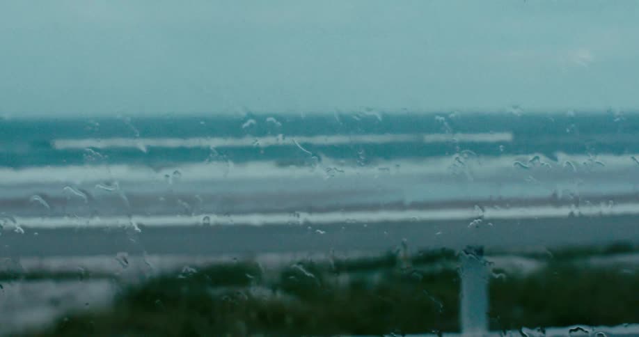 A tranquil yet melancholic view of the ocean as raindrops slowly trickle down a windowpane