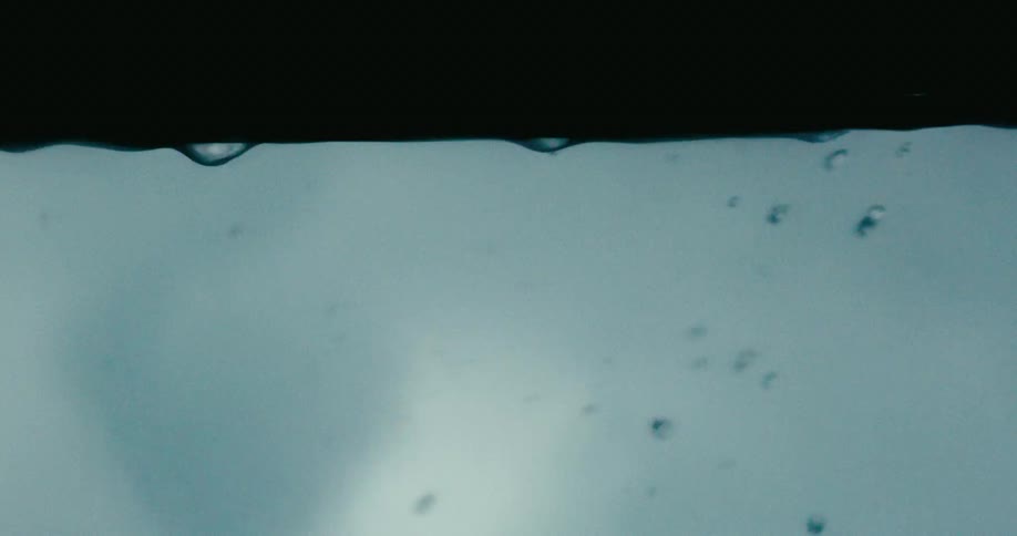 An abstract close-up captures raindrops forming and falling in slow motion against a gloomy, overcast sky