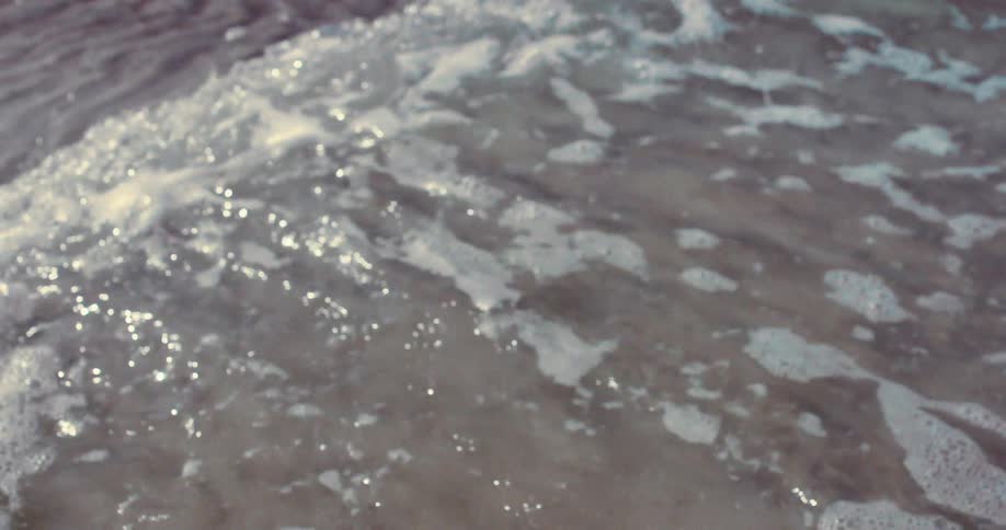 A soft-focus, slow-motion close-up of a gentle ocean wave with white foam washing over the rippled sand on the shore