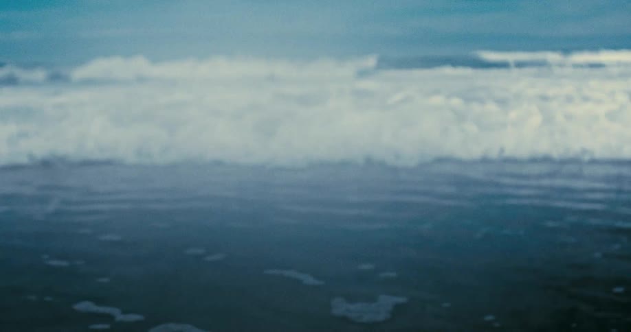 An out-of-focus ocean wave slowly rolls towards the shore and crashes into white foam in a serene, slow-motion shot