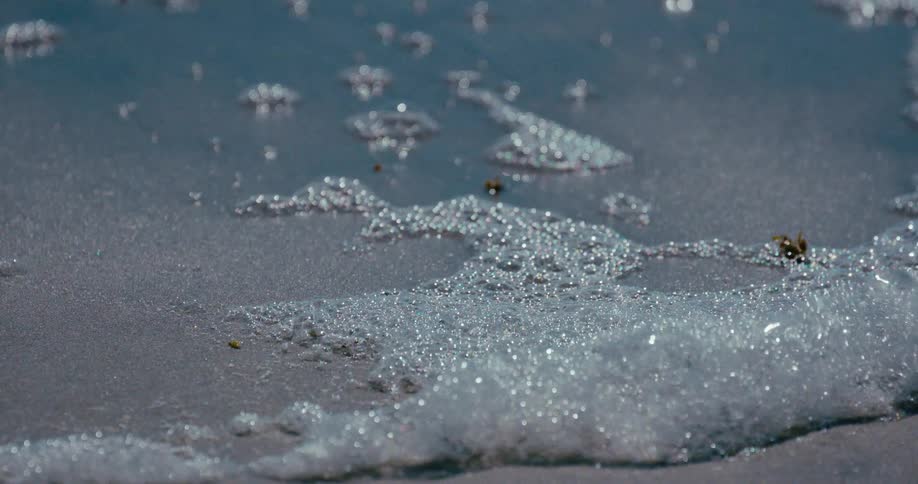 A mesmerizing slow-motion close-up of sparkling seafoam bubbles gently lapping on a wet sandy shore