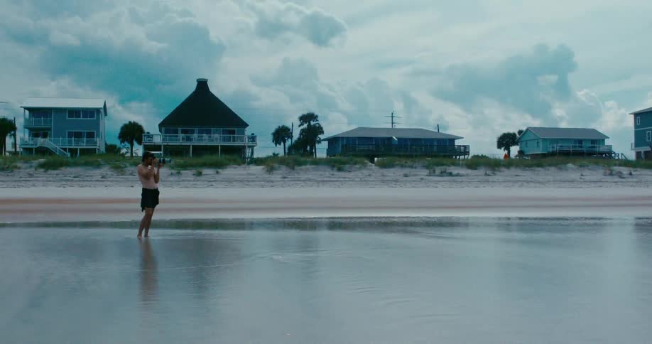 A lone photographer stands in the shallow ocean tide, capturing the view of beach houses under a vast, cloudy sky