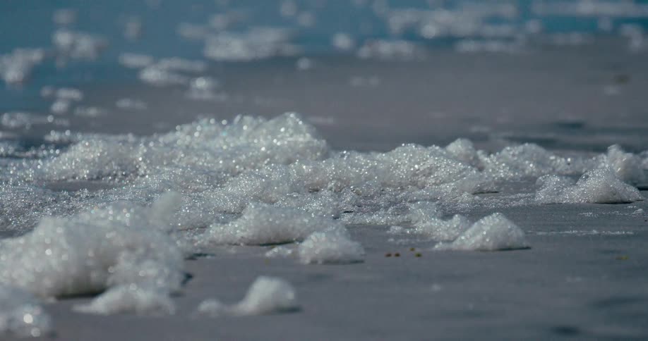 Glistening sea foam is blown across a sandy beach in slow motion