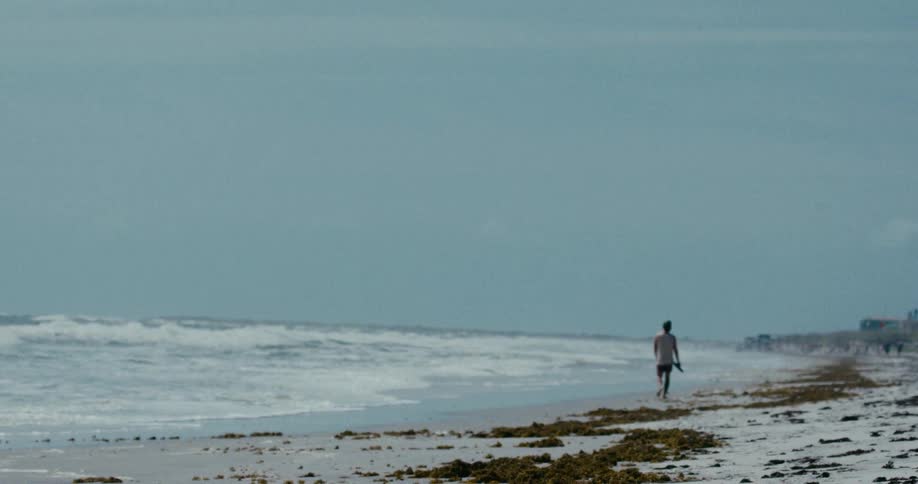 A soft focus shot shows a solitary man walking along a sandy beach as ocean waves crash on the shore