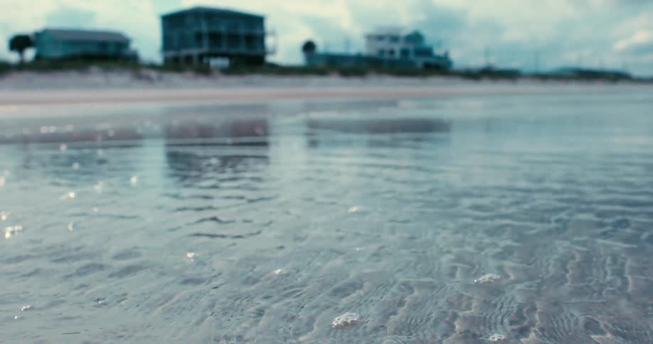 Shallow ocean water gently recedes from the sandy shore, creating tranquil ripples with beachfront houses blurred in the background