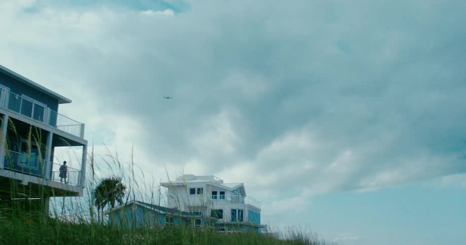 A small airplane flies slowly across a vast, cloudy sky over modern coastal beach houses nestled in grassy dunes