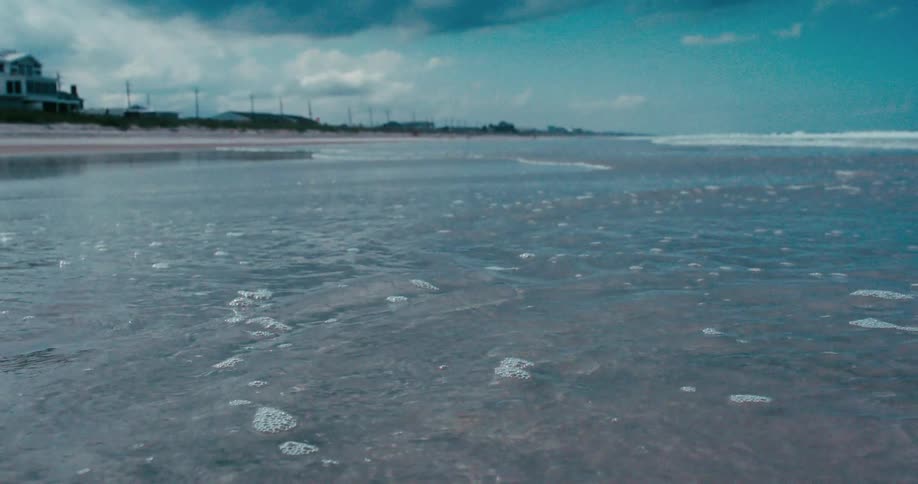 Gentle ocean water with bubbles recedes from the shore in a calming, low-angle, slow-motion shot