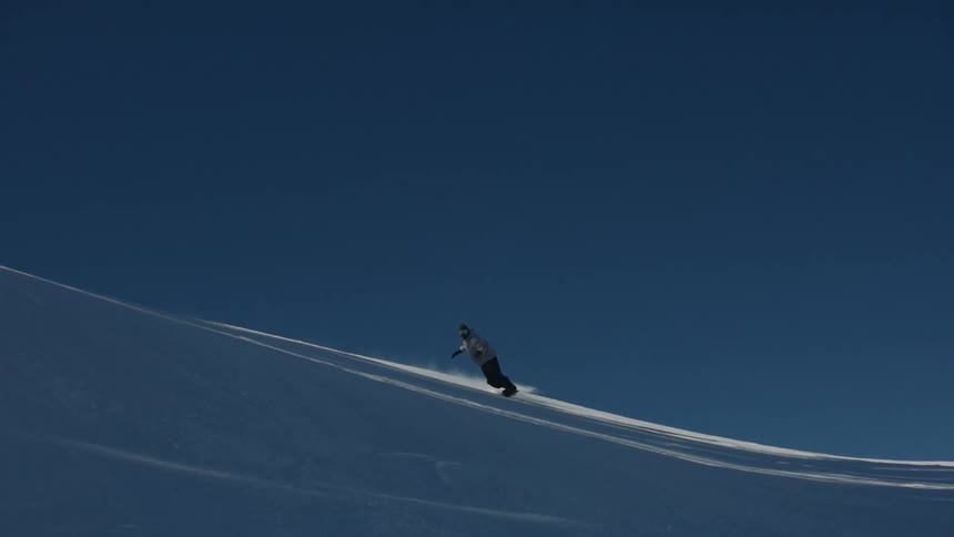 A lone snowboarder carves down a pristine, untouched snowy mountain under a vast, deep blue sky in this cinemagraph