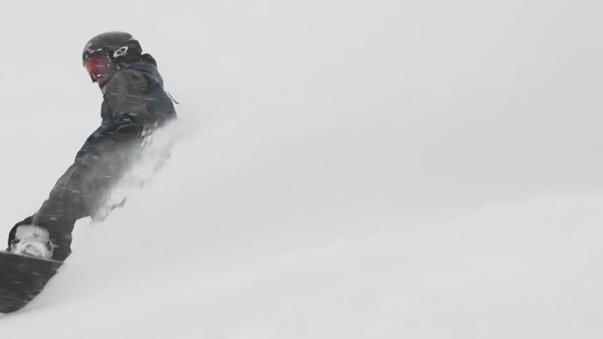 A snowboarder carves through deep powder snow in slow motion, kicking up a huge spray and disappearing from the frame