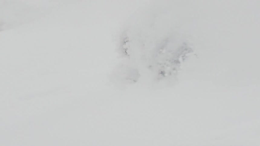 A snowboarder carves through deep powder snow in slow motion as heavy snow falls around them on a mountain