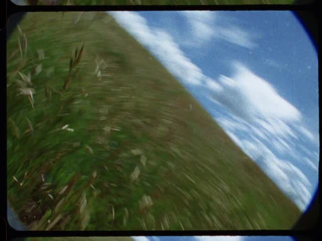 A dizzying first-person perspective shot on vintage film tumbles and runs through a tall grassy field under a blue sky with clouds