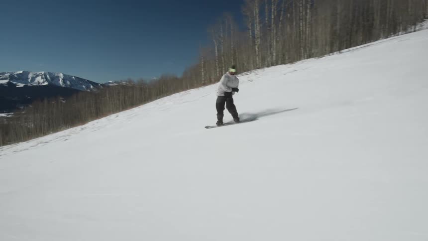 A snowboarder carves down a groomed mountain slope in slow motion on a beautiful sunny day