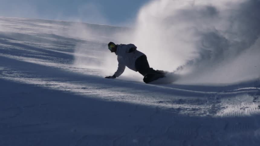 A snowboarder carves down a snowy slope, kicking up a huge spray of fresh powder in epic slow motion