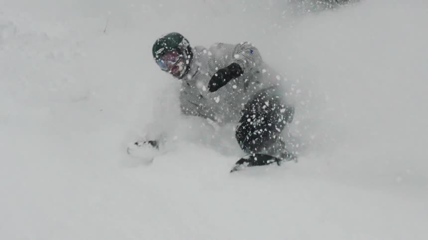 A snowboarder in a helmet carves through deep powder, kicking up a huge spray of snow in super slow motion