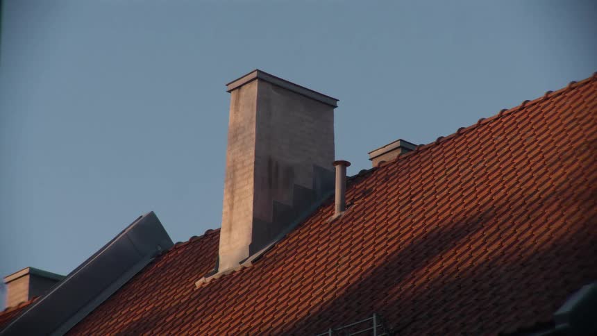 A close-up of a brick chimney on a red tiled roof basking in the warm glow of the setting sun against a clear blue sky