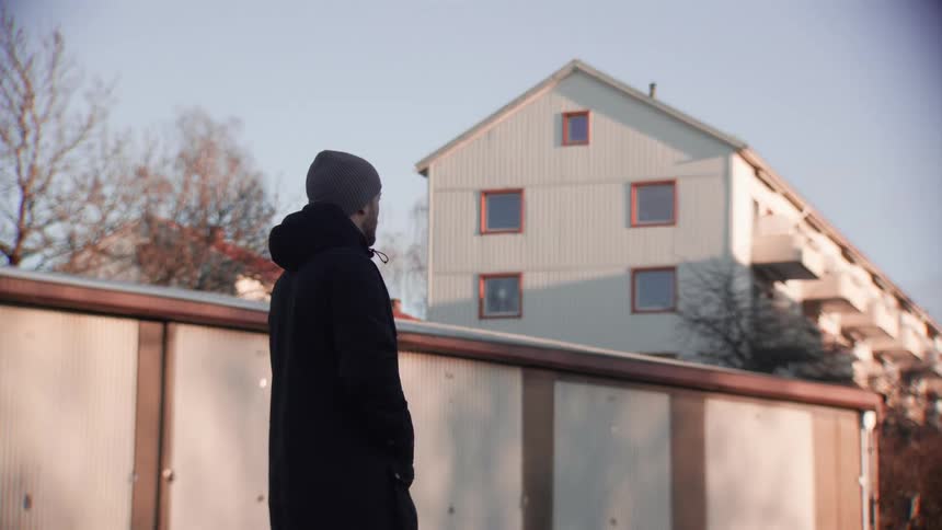 A man in a winter coat and beanie stands and gazes at an apartment building on a sunny day
