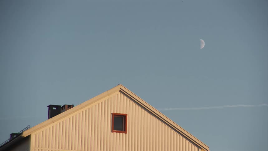 A half moon is visible in the daytime sky above the peak of a residential house