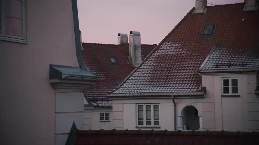 A peaceful shot shows snow-covered tiled rooftops of old buildings against a soft pink sky at sunset
