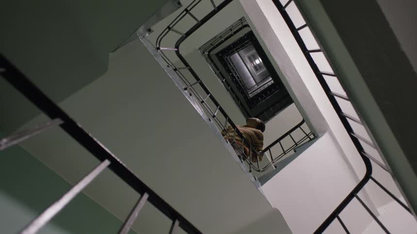 An upward-facing view captures a person slowly walking up the many flights of a square spiral stairwell