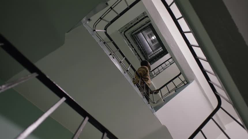 A woman walks down an endless flight of square spiral stairs as seen from a low angle looking up