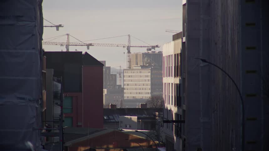 A static shot between two buildings under renovation reveals a cityscape with multiple construction cranes in the distance