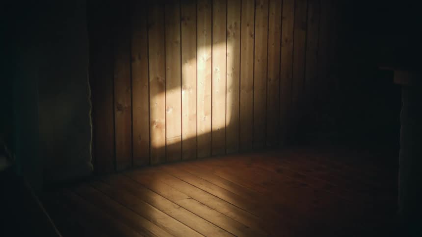 A single beam of sunlight illuminates the wooden wall and floor of a dark, empty, rustic room