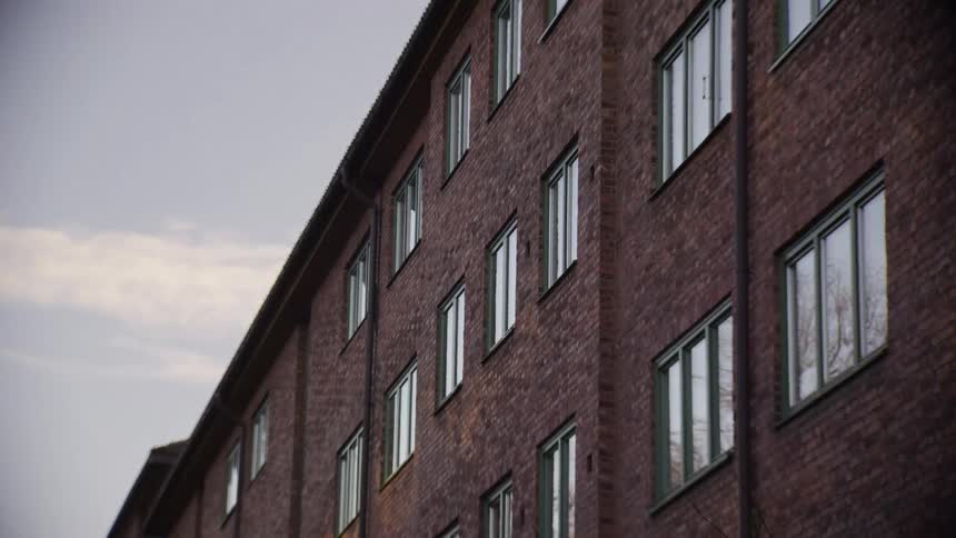 A low-angle static shot of a red brick apartment building with many windows under an overcast sky
