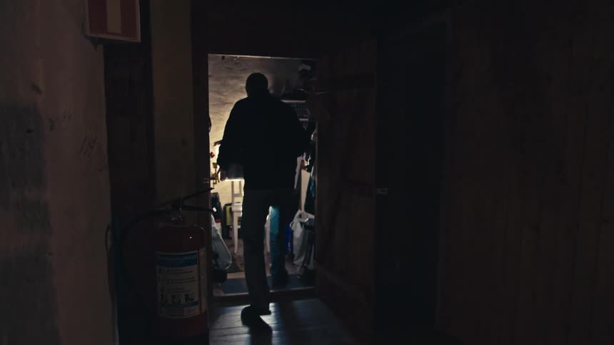 A man carries a plastic container through a dark, rustic wooden hallway into a cluttered storage room
