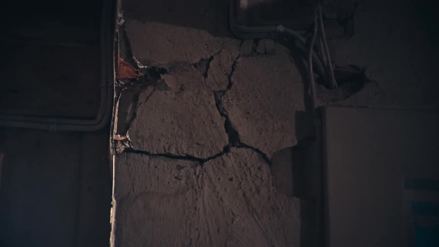 A beam of light illuminates deep cracks in a crumbling concrete wall inside a dark, derelict building