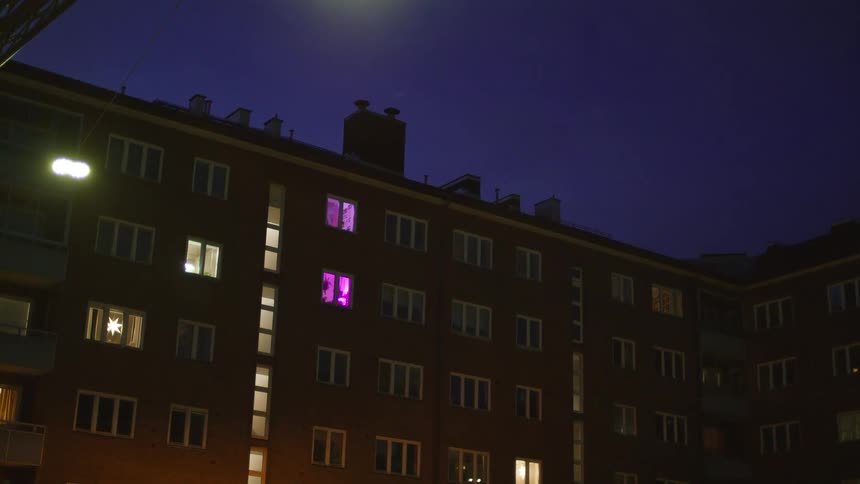 A low-angle shot of a city apartment building at night with a purple light glowing from one of the windows