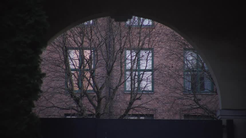 A slow zoom-out reveals a view of a red brick building and bare trees through a dark, mysterious archway at dusk