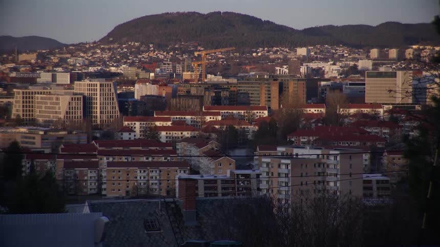 A slow timelapse captures the afternoon sun moving across a cityscape with modern and classic architecture backed by a large forested hill