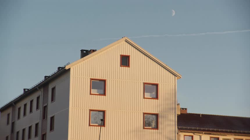 A daytime view of a white apartment building with a crescent moon and a contrail in the clear blue sky