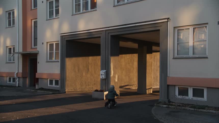 A small child wearing a helmet rides a balance bike across a courtyard and into a building breezeway at sunset