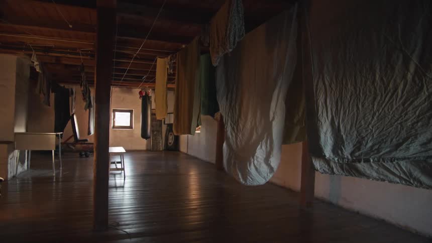 A slow pull-out shot reveals a dimly lit, rustic laundry room with clothes and sheets hanging on lines to dry