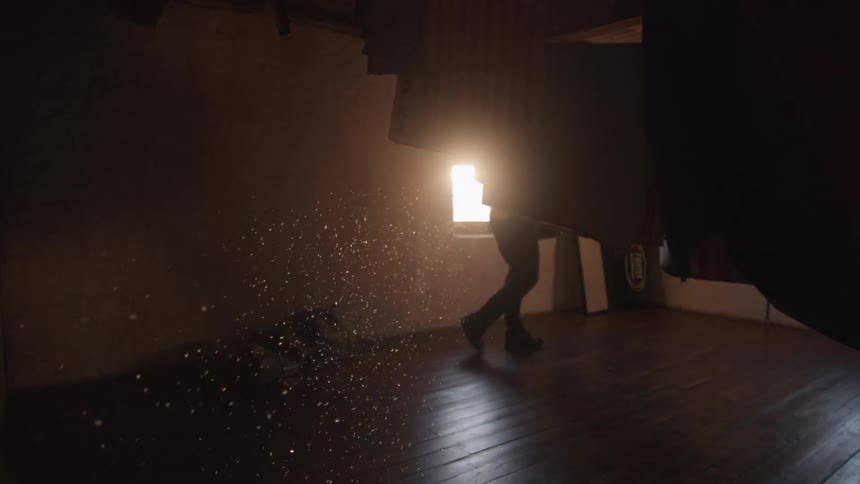 Atmospheric slow-motion shot of a man stirring up dust and wood shavings that glitter in a single beam of light from a window in a dark room