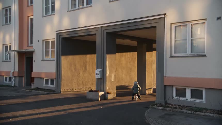 A small toddler wearing a helmet rides a balance bike through an apartment courtyard during golden hour