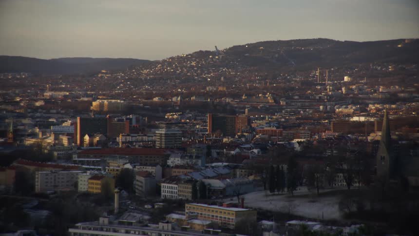 A wide shot of the city of Oslo, Norway, during a calm winter sunset with the Holmenkollen ski jump visible on the hill in the background
