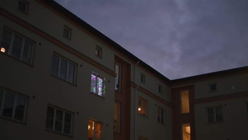 Colorful flashing party lights illuminate a single apartment window against a dark, cloudy sky at dusk