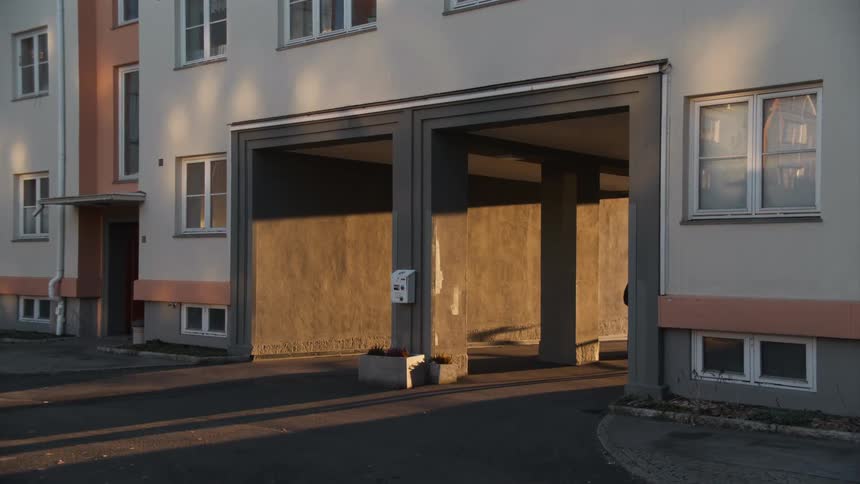 A man in a black coat walks into the golden hour shadows of an apartment building archway