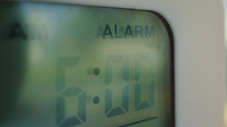 A digital alarm clock face flashes 6:00 AM as the morning wake-up alarm goes off