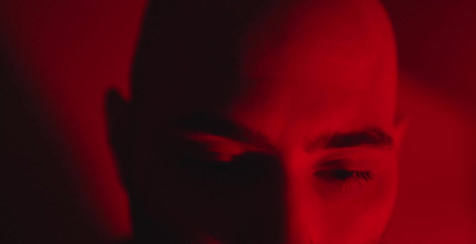 A blurry close-up of a bald man's face slowly comes into focus, illuminated by a dramatic red light that casts deep shadows