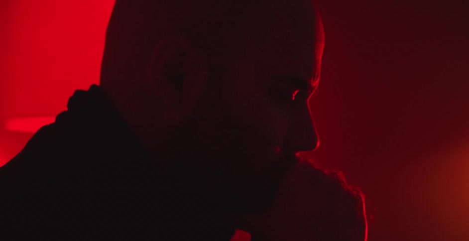 A serious man sits in deep thought, his face silhouetted by intense red lighting